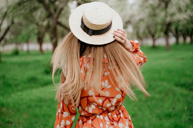 Mulher loura bonita nova no jardim de florescência. Árvores de primavera em flor. Vestido laranja e chapéu de palha.