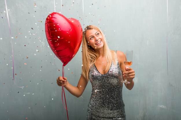 Mulher loura nova elegante que comemora um evento, guardando um copo de champanhe e um balão vermelho do coração.