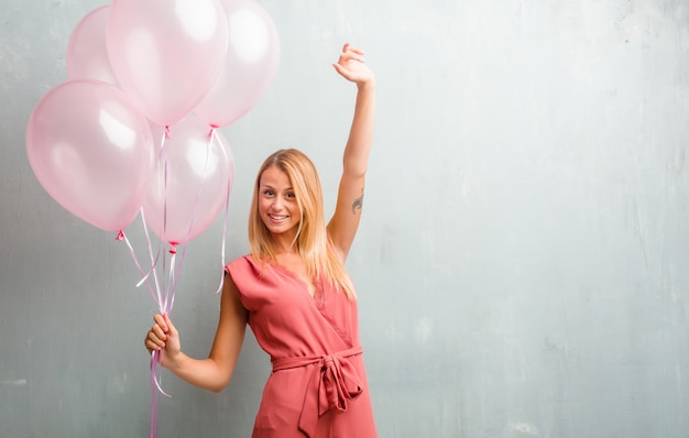 Mulher loura nova elegante que mantém balões cor-de-rosa contra uma parede.