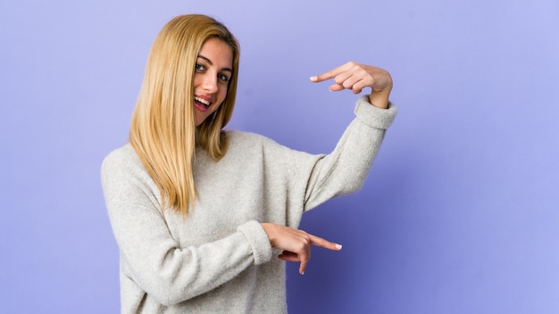 Mulher loura nova isolada no roxo que guarda algo pequeno com o dedo indicador, sorrindo e seguro.