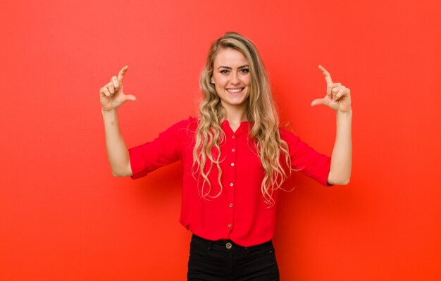 Mulher loura nova que quadro ou que descreve o próprio sorriso com ambas as mãos, olhando positivo e feliz, conceito do bem-estar na parede vermelha