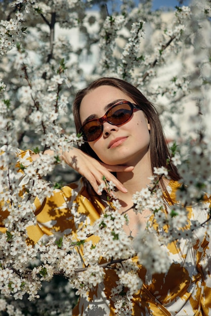 Mulher macia em óculos de sol com olhos fechados, aproveitando a primavera perto de uma árvore florescente com flores