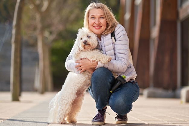 Mulher madura andando com cachorro branco fofo na cidade de verão.