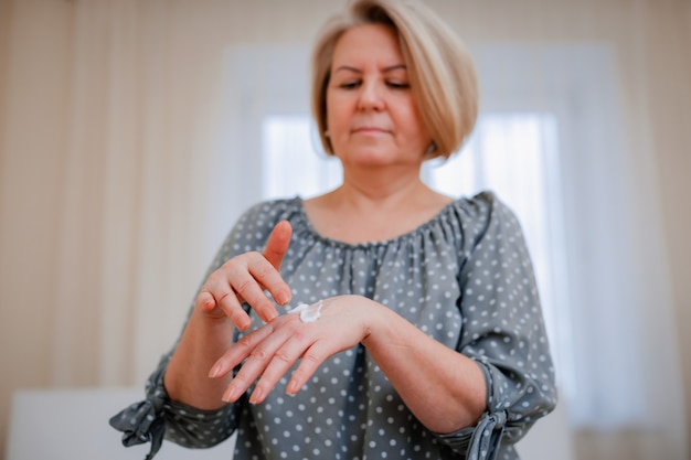 Mulher madura aplica um creme cosmético hidratante antienvelhecimento às mãos, sorri para uma senhora de meia-idade com um cuidado de pele limpo e macio