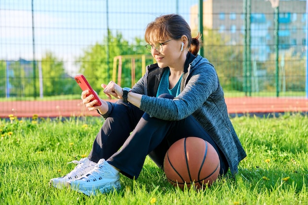 Mulher madura ativa com bola de basquete e smartphone ao ar livre