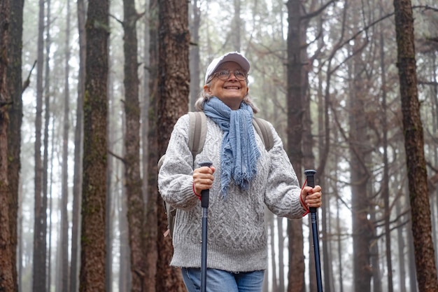 Mulher madura ativa e feliz com mochila caminhando na floresta de montanha em um dia nebuloso com a ajuda de postes