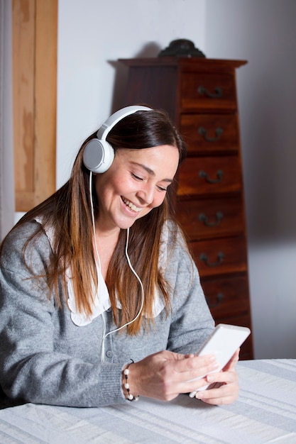 Foto mulher madura atraente em casa usando celular, vestindo um suéter cinza e fones de ouvido, videoconferência