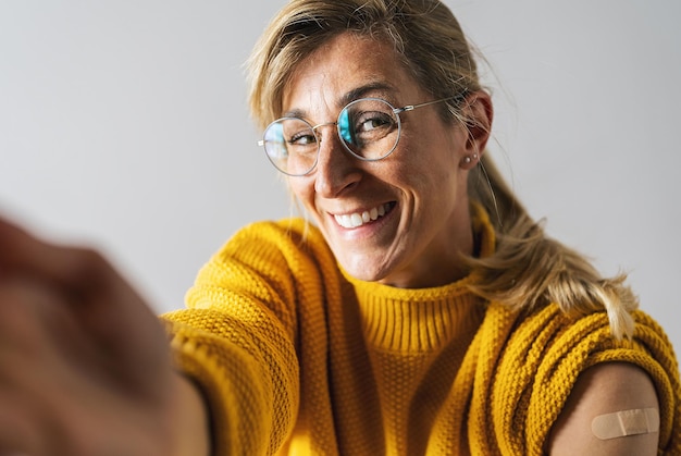 Mulher madura atraente sorrindo depois de receber uma vacina corona Mulher segurando a manga da camisa e mostrando o braço com curativo depois de receber a vacinação esticando o braço para a câmera para uma selfie