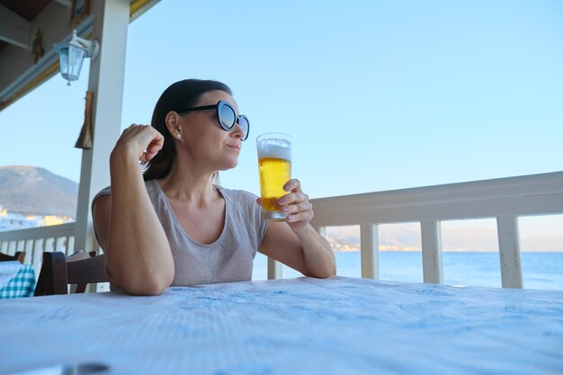 Mulher madura bonita descansando sentado em um café à beira-mar bebendo um copo de cerveja gelada