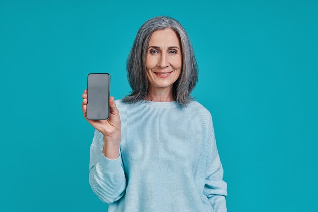 Mulher madura bonita mostrando um telefone inteligente e sorrindo em pé contra um fundo azul