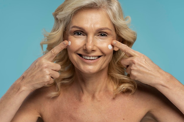 Mulher madura caucasiana sorridente dos anos 50 aplicando creme facial antienvelhecimento isolado sobre fundo azul