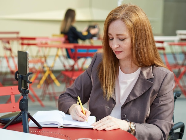 Mulher madura closeup que usa uma cadeira de rodas usando smartphone para trabalho de aprendizado remoto fazendo anotações no caderno