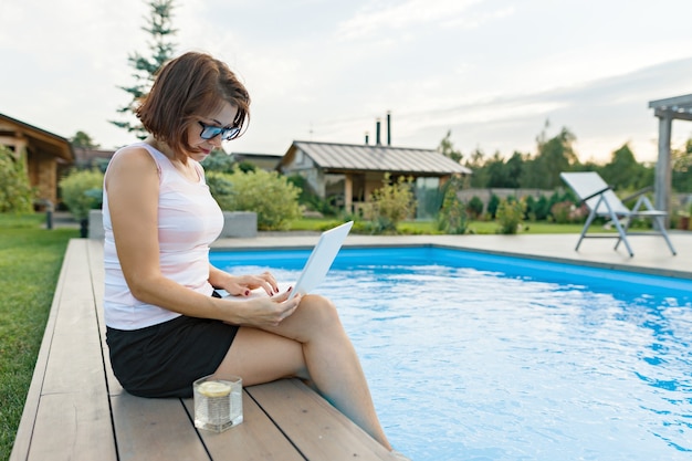 Mulher madura com laptop perto de piscina particular