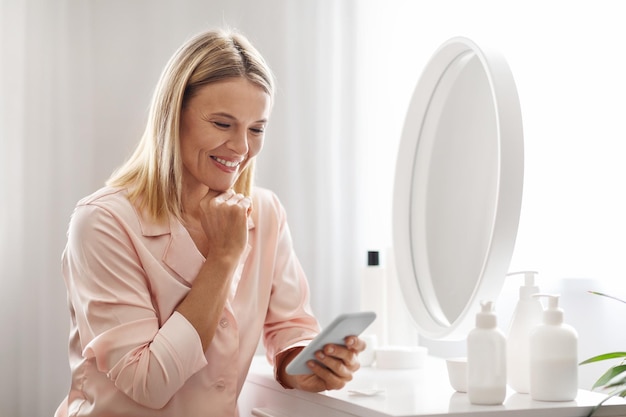 Mulher madura com smartphone lendo truques de beleza enquanto se prepara em casa