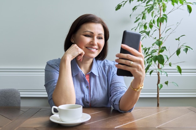 Mulher madura com smartphone sentado em casa sorrindo olhando para a tela do telefone