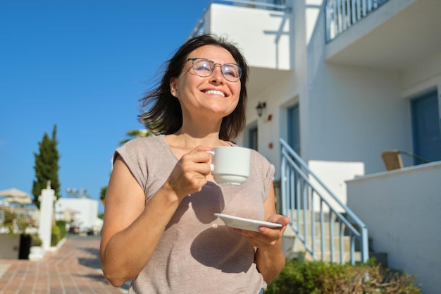 Mulher madura de óculos com uma xícara de café caminhando ao ar livre, retrato de mulher de meia-idade sorridente