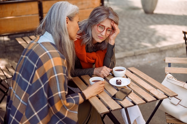 Mulher madura deprimida com óculos e amiga cuidadosa si em um café de rua no dia de outono