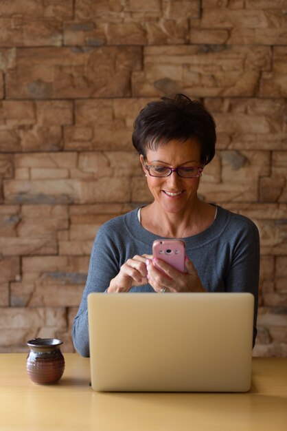 Mulher madura e feliz sorrindo enquanto usa o telefone celular com o laptop na mesa de madeira contra a parede de tijolos