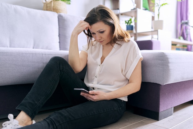 Mulher madura e triste lendo smartphone em casa sentado no chão.