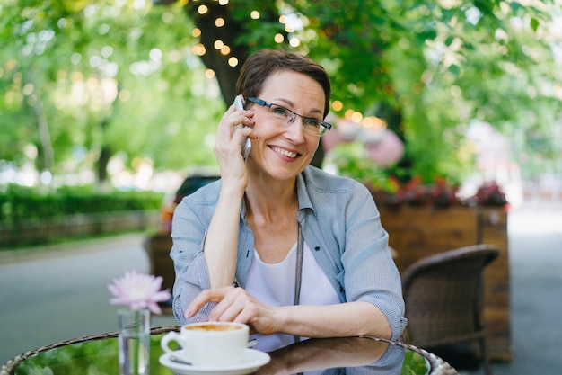 Mulher madura em óculos falando no celular no café ao ar livre ao ar livre