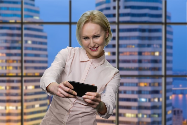 Mulher madura está jogando em seu smartphone. secretária atraente caucasiana em blusa branca. vista da cidade à noite em segundo plano.