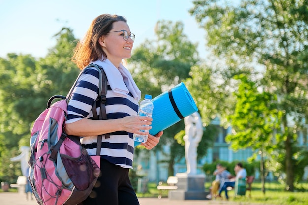 Mulher madura feliz ao ar livre andando após exercícios fit no parque, com mochila, tapete de ioga. Esportes ativos, estilo de vida saudável de pessoas de meia-idade, corpo e beleza