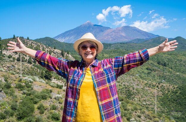 Mulher madura feliz com chapéu de palha em pé ao ar livre, desfrutando de caminhada. Montanhas com o vulcão Teide ao fundo