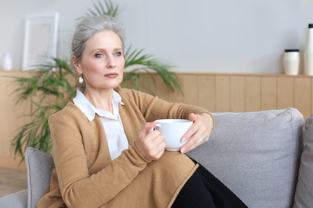 Mulher madura feliz descansando em um sofá confortável beber café ou chá, olhando para longe, relaxando no sofá aconchegante em casa, desfrutar de uma bebida quente