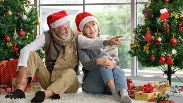 Mulher madura feliz e homem idoso examinando o presente enquanto está sentado no tapete entre árvores de Natal decoradas contra a janela de casa