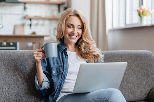 Mulher madura feliz e relaxada descansando no sofá vendo filmes filmes webinars vídeo no laptop bebendo café quente chá bebida em casa compras on-line aprendizagem à distância