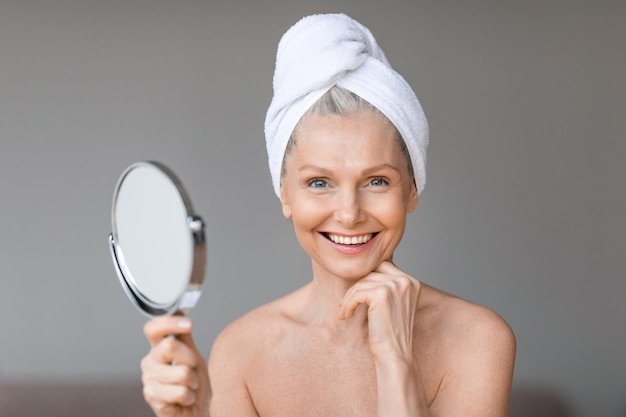 Mulher madura feliz fazendo rotina matinal depois do banho, segurando o espelho e sorrindo para o quarto da câmera