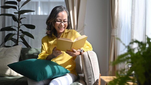 Mulher madura feliz lendo livro enquanto descansava confortável na sala de estar brilhante