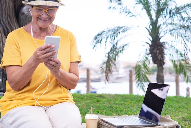 Mulher madura feliz sentada ao ar livre usando um computador laptop com smartphone ao seu lado