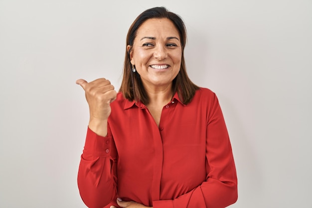 Mulher madura hispânica em pé sobre fundo branco sorrindo com cara de feliz olhando e apontando para o lado com o polegar para cima.