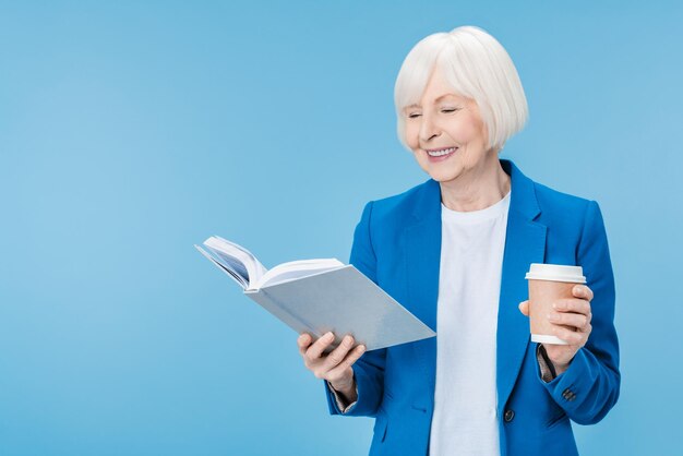 Mulher madura lendo livro com copo de bebida sobre fundo azul