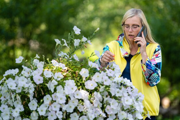 Mulher madura loira, brilhantemente vestida, falando no smartphone no conceito de comunicação on-line do jardim