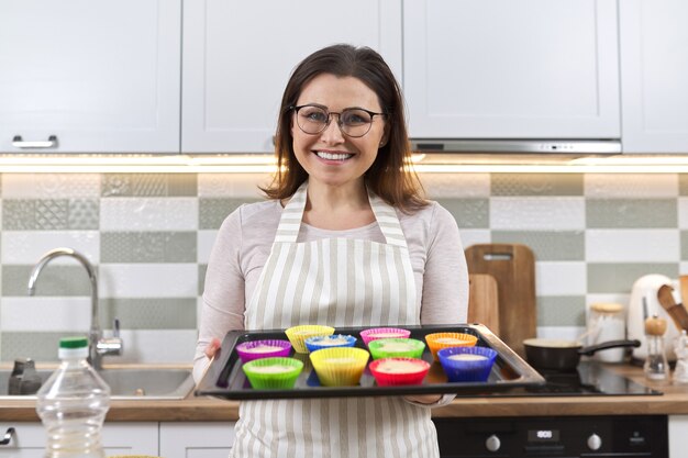 Mulher madura no avental com assadeira com cupcakes sendo preparados. mulher em casa cozinhando, assados caseiros, comida saudável, hobby