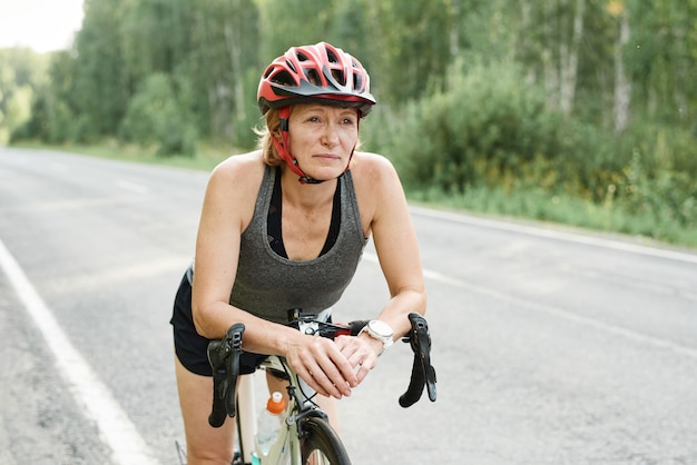 Mulher madura no capacete, sentada na bicicleta, participando de uma competição esportiva ao ar livre