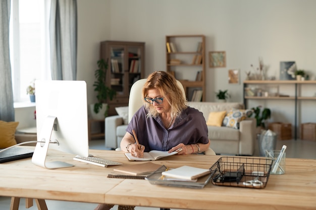 Mulher madura ocupada usando óculos e roupas casuais, fazendo anotações ou anotando pontos do plano de trabalho na frente do monitor do computador