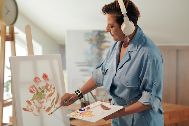 Foto mulher madura ouvindo música em fones de ouvido ao pintar em cavalete