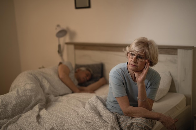 Mulher madura pensativa se sentindo sem dormir e sentada na cama enquanto o marido está dormindo no fundo