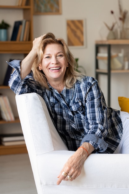 Foto mulher madura rindo em roupas casuais olhando para você com um sorriso cheio de dentes enquanto está sentado em uma poltrona branca em frente à câmera na sala de estar