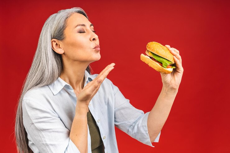 Foto mulher madura sênior comendo hambúrguer com satisfação a avó gosta de saboroso hambúrguer para viagem deliciosa mordida de entrega de fastfood de pedido de hambúrguer enquanto está com fome isolado sobre fundo vermelho