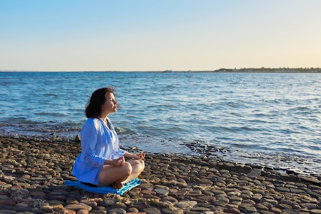 Mulher madura sentada em posição de lótus meditando na praia