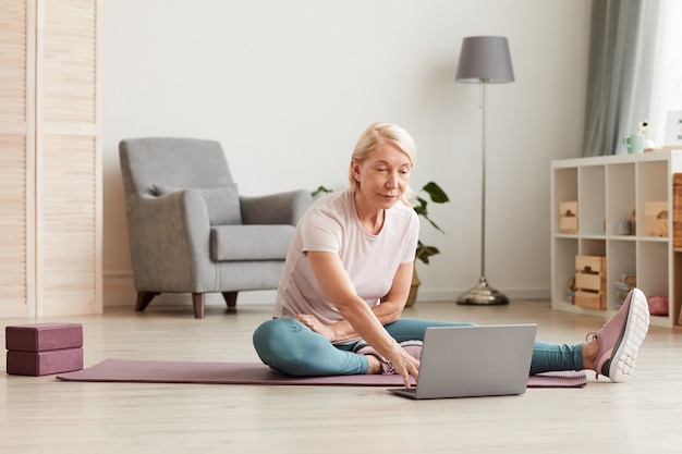 Mulher madura sentada no chão usando um laptop durante o treinamento esportivo em casa