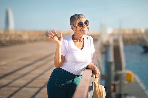 Mulher madura sorridente acenando com a mão posando no cais com veleiros