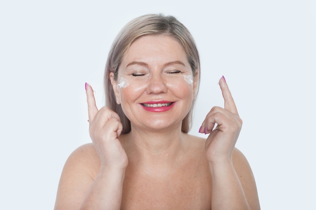Mulher madura sorridente aplicando creme rejuvenescedor em seu retrato de pele de rosto em fundo branco