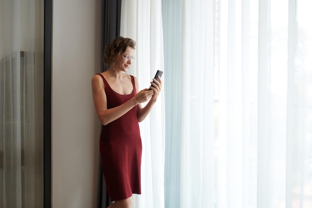 Mulher madura sorridente de vestido vermelho profundo em pé na janela e lendo mensagens de texto ou notificações