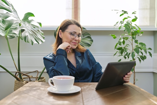 Mulher madura sorridente tomando café da manhã em casa de pijama com uma xícara de chá, sentado à mesa lendo o tablet digital. Mulher feliz de ler e-mails, notícias. Emoções positivas, informação, vida, pessoas dos anos 40