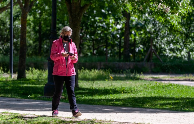 Mulher madura treina caminhando por um parque enquanto consulta redes sociais em seu telefone celular